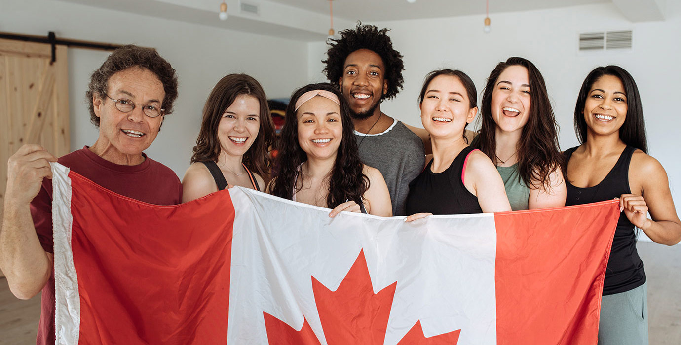A happy group of multi-ethnic people stand behind a large Canada flag.