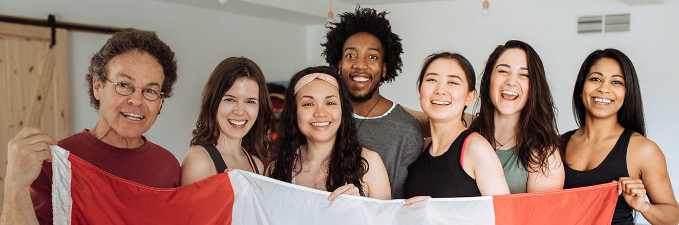 A happy group of multi-ethnic people stand behind a large Canada flag.