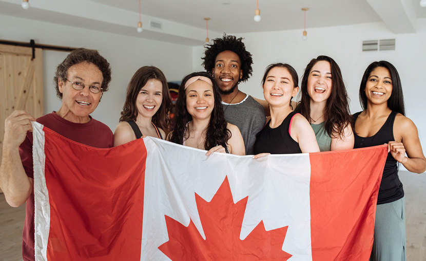 A happy group of multi-ethnic people stand behind a large Canada flag.