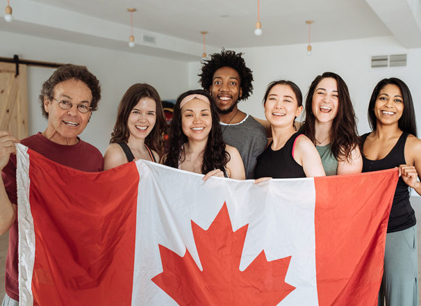 A happy group of multi-ethnic people stand behind a large Canada flag.