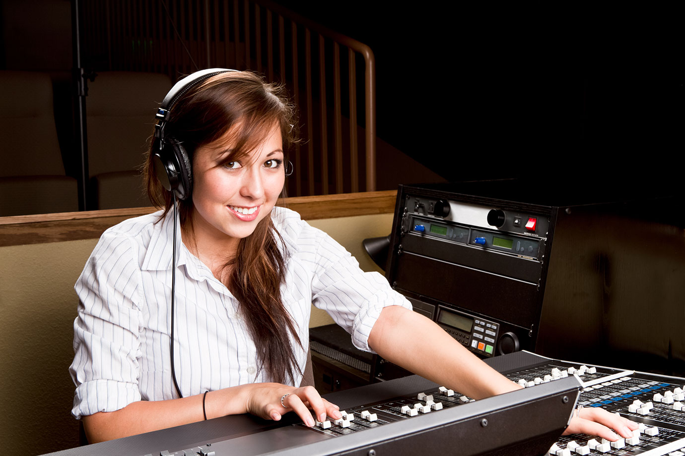 A person wearing headphones sits in front of a mixing console while smiling.