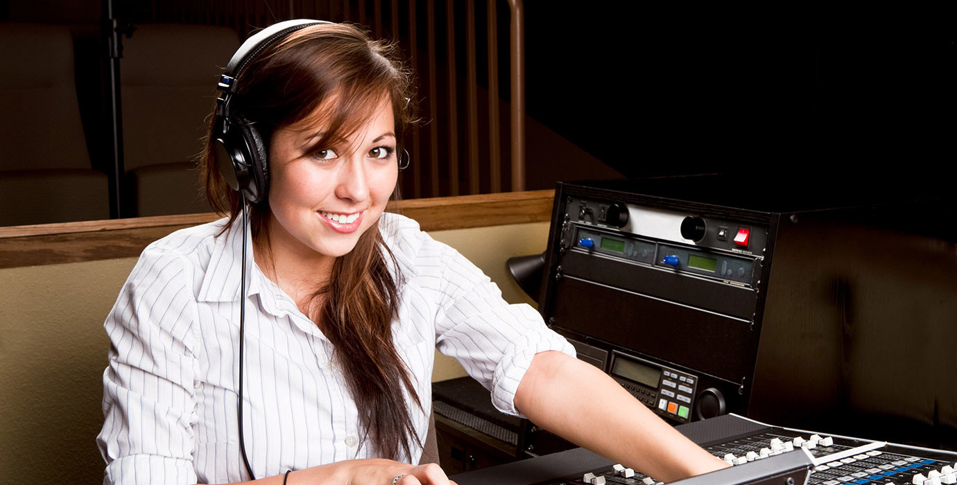 A person wearing headphones sits in front of a mixing console while smiling.