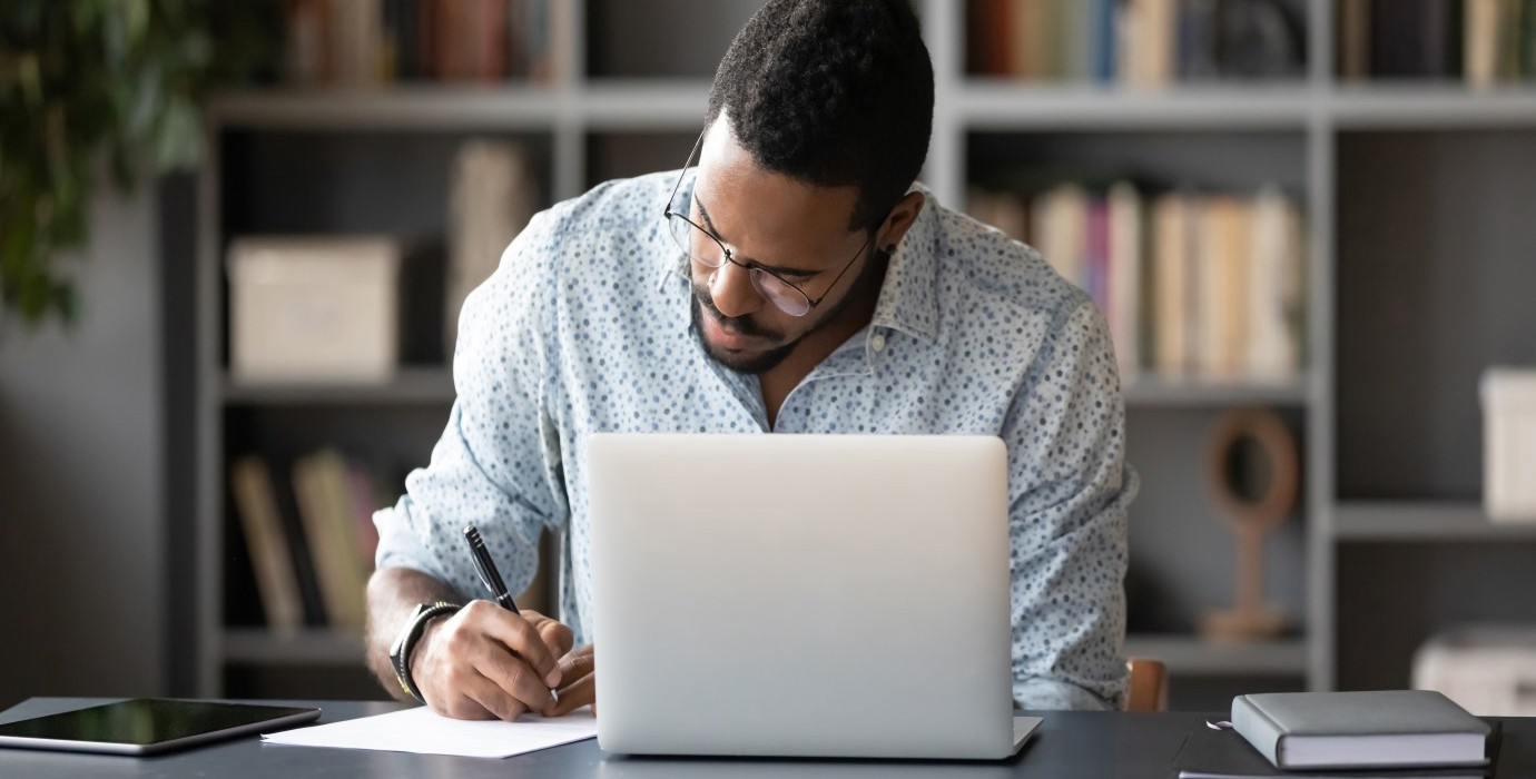 Student taking notes while using laptop at home office desk.
