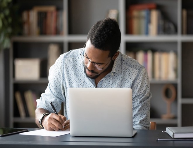 Student taking notes while using laptop at home office desk.