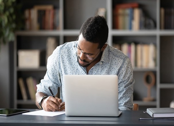 Student taking notes while using laptop at home office desk.