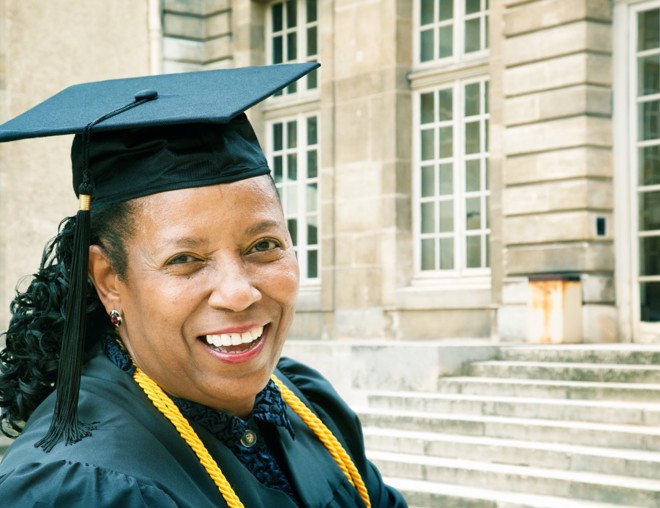 Woman in a graduation cap and gown smiling at the camera outside of a school.