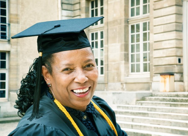 Woman in a graduation cap and gown smiling at the camera outside of a school.
