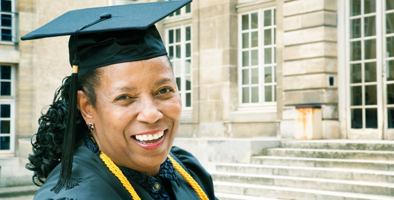 Woman in a graduation cap and gown smiling at the camera outside of a school.