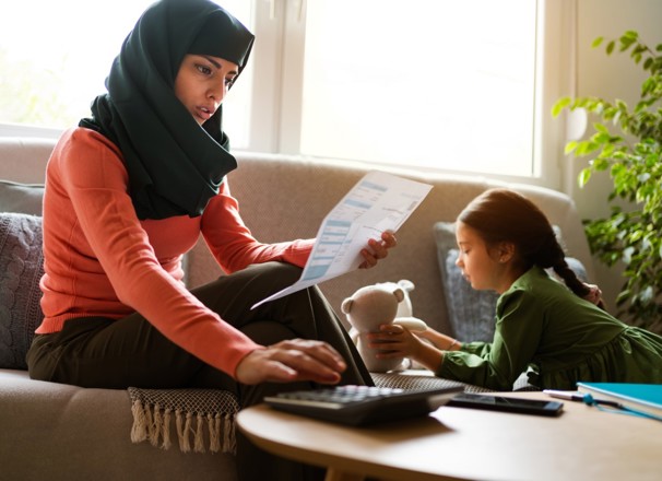 Mother paying bills while sitting on the couch with her young daughter, who is playing with a stuffed animal.