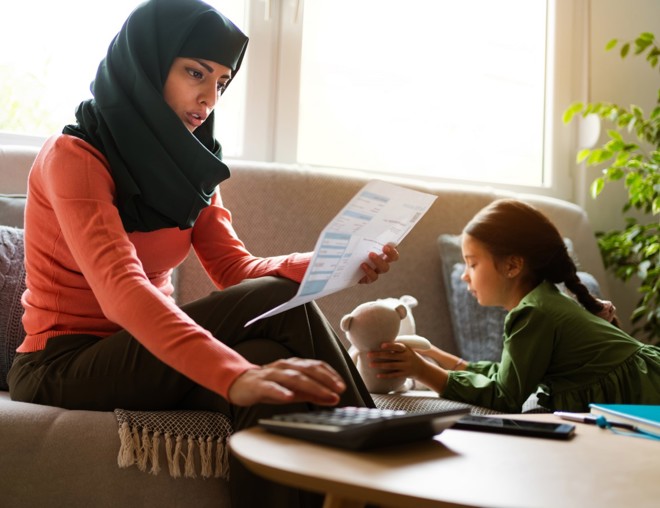 Mother paying bills while sitting on the couch with her young daughter, who is playing with a stuffed animal.