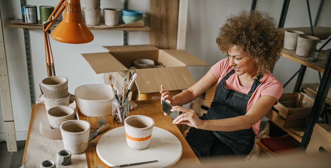 Woman wearing a denim apron taking a picture of a clay pot she painted