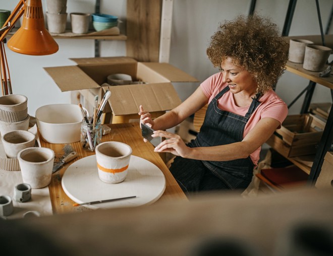 Woman wearing a denim apron taking a picture of a clay pot she painted