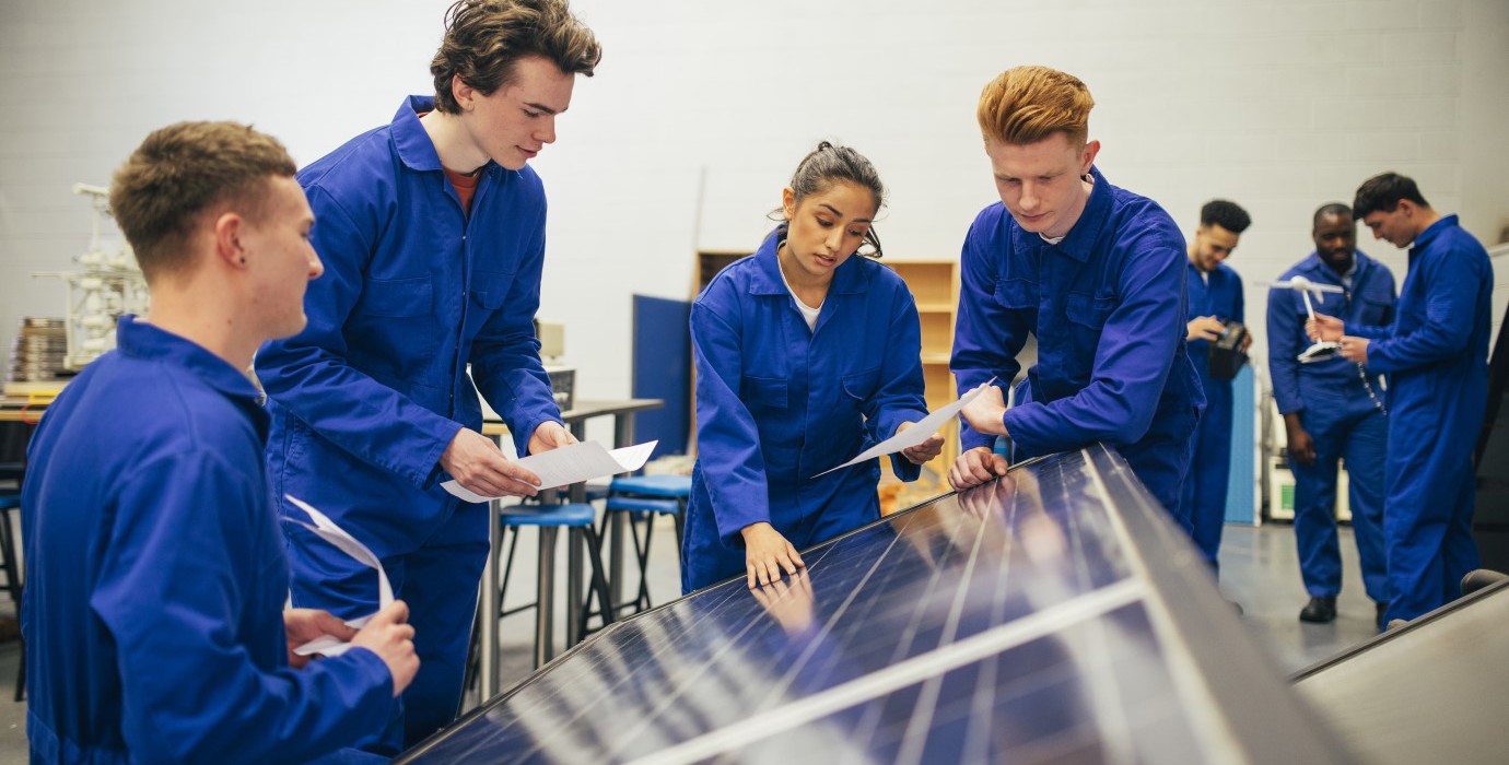 Four students look over a solar panel and make notes.