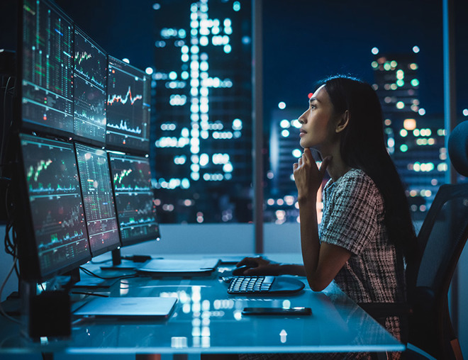 analyst at computer at night looking at market charts on her multi-monitor workstation