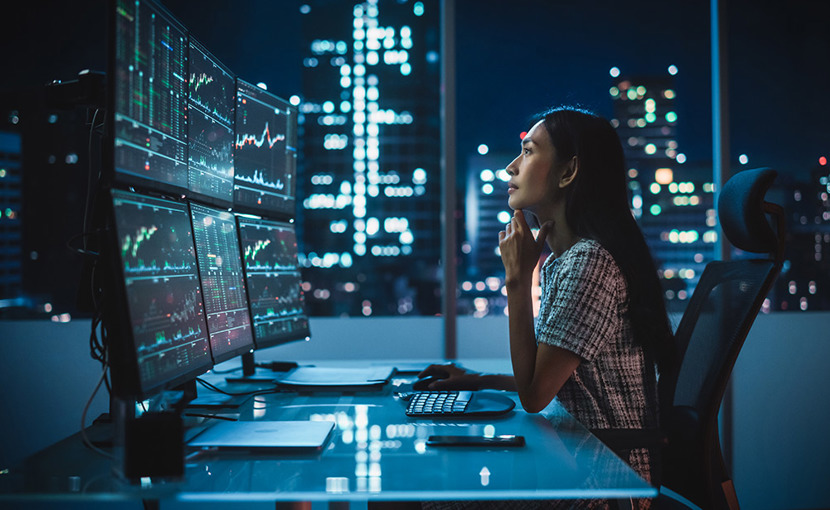 analyst at computer at night looking at market charts on her multi-monitor workstation