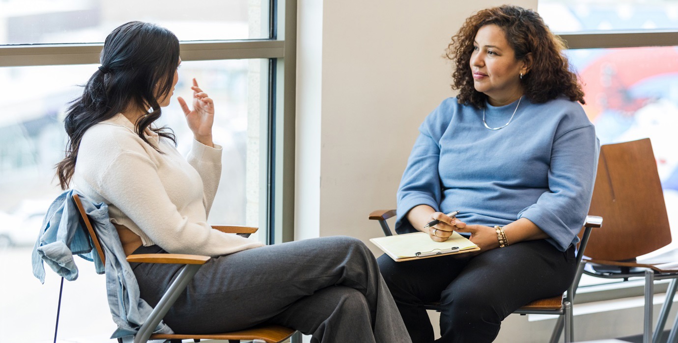 Two people talking at a table by a window.