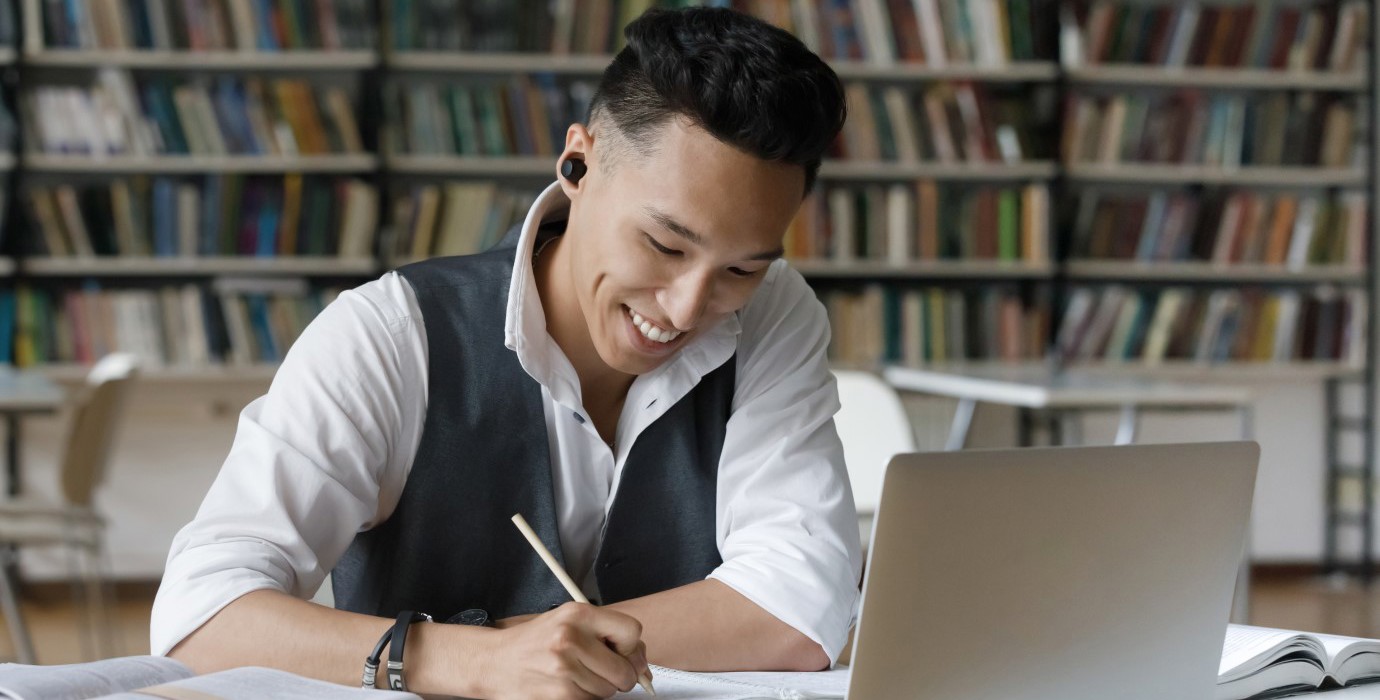 Student taking notes while using laptop in library.