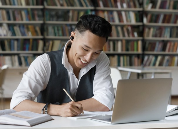 Student taking notes while using laptop in library.
