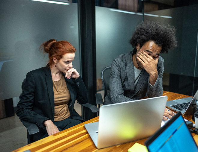 2 colleagues at desk in front of laptop looking frustrated
