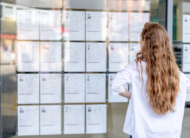 A person with long hair stands in front of a glass window, carefully reviewing job listings that are displayed on the wall outside a public space.