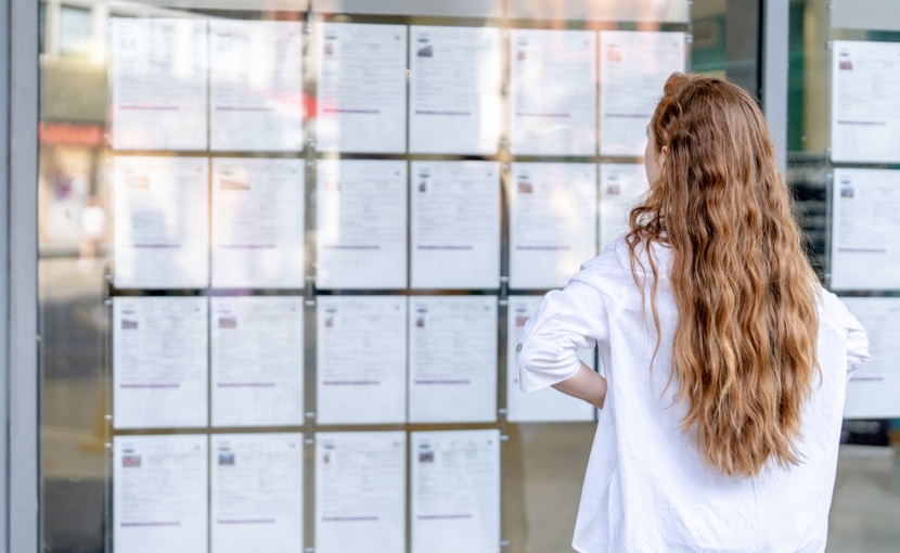 A person with long hair stands in front of a glass window, carefully reviewing job listings that are displayed on the wall outside a public space.