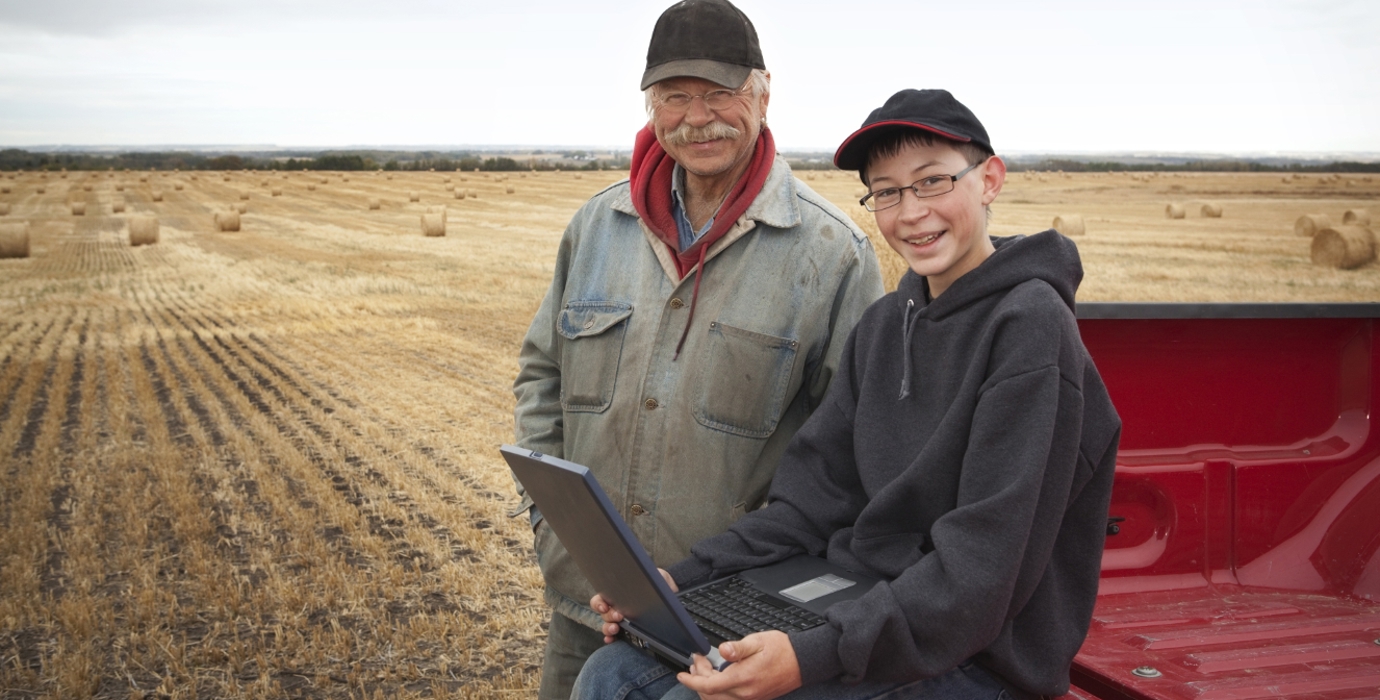 Father and son in a hay field looking at a laptop while sitting on the bed of a pickup truck.