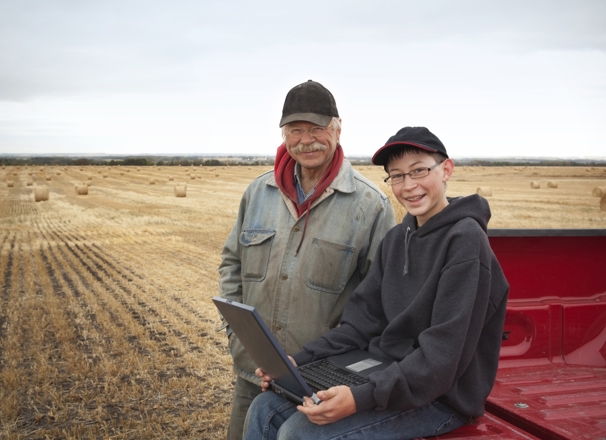 Father and son in a hay field looking at a laptop while sitting on the bed of a pickup truck.