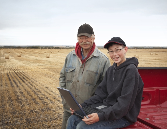 Father and son in a hay field looking at a laptop while sitting on the bed of a pickup truck.